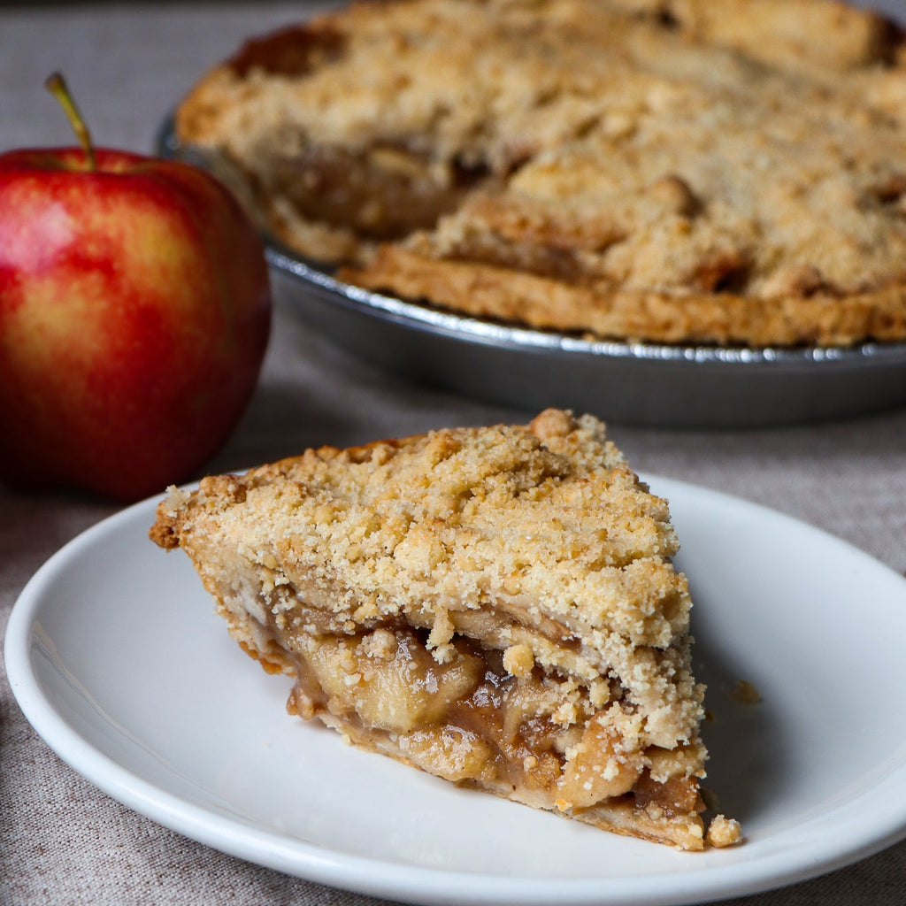 Slice of apple pie on a plate with a whole pie and an apple in the background.