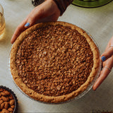 Person holding a black bottom oat pie on a table with almonds and glasses.