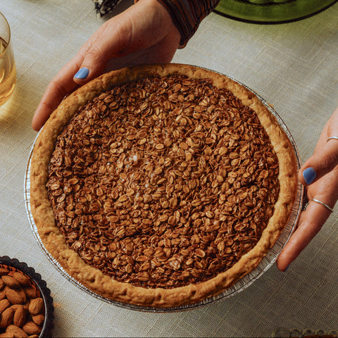 Person holding a black bottom oat pie on a table with almonds and glasses.