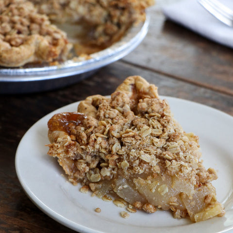 Slice of pear pie with crumble topping on a white plate, with the rest of the pie in the background.