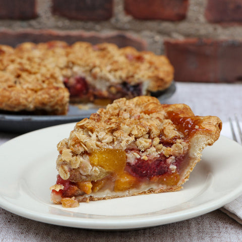 Slice of  stonefruit pie with crumble topping on a white plate against a brick wall background