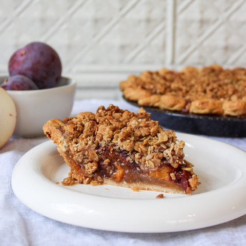 Slice of plum and peach pie on a white plate with a whole pie and plums in the background.
